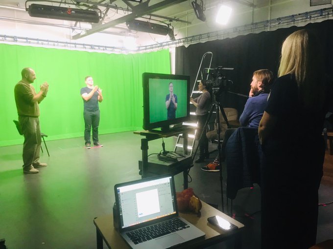 Image of a recording studio with a green screen in the background. Three team members are discussing a sign. The signer is visible on the video recorded and the videographer is watching on.