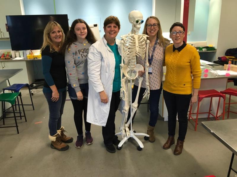 Image of five women, some wearing labcoats, standing around a skeleton in a science lab