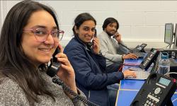 Three students (two female & one male) holding land phones to thier ears with computers in front of them