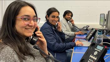 Three students (two female & one male) holding land phones to thier ears with computers in front of them