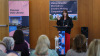 Shows a woman speaking from a lectern to a seated audience