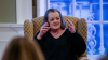 Shows a woman sitting in a chair, gesturing with her hands