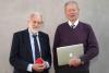 Two men in suits posing in front of the camera one is holding his laptop, they look serious