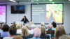 A woman standing at a lectern speaking to a seated audience. The screen behind her reads Welcome to the DCU Library Creative Writing Competition awards 2025