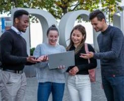 A group of different students of all backgrounds are all standing in front of a DCU Sign. They are all looking at and talking in front of a laptop, as thought they are working on a project together.