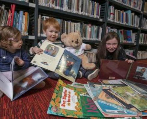 Three children sit in book shelves with books sprawled on the floor in front of them