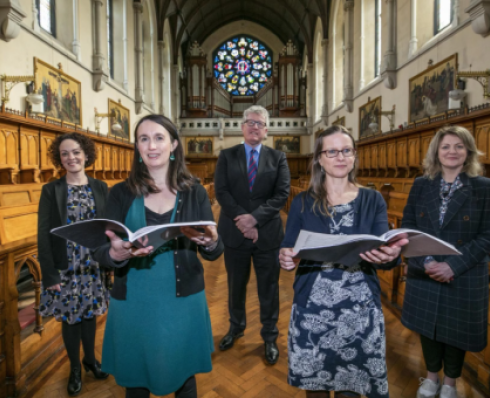 Five people stand in church aisle. The two who stand in front are holding song books