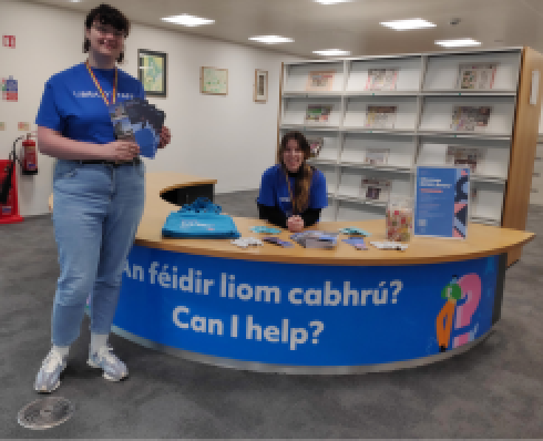 Two people in blue tshirts beside a desk with the text: An féidir liom cabhrú? Can I help?