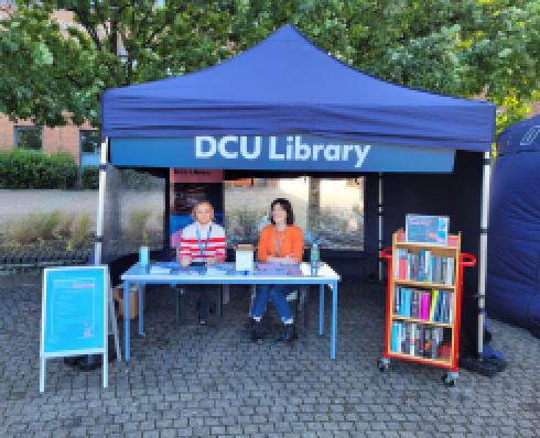 A blue topped tent with a sign above saying DCU Library. In the tent are 2 friendly library staff behind a desk, with a trolley of books and a sign listing library orientation activities