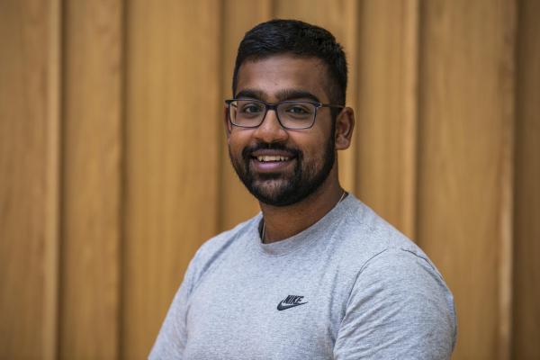 A professional portrait of a man with brown skin, glasses, black hair, a beard and moustache. He is wearing a grey shirt and smiling to the camera. 