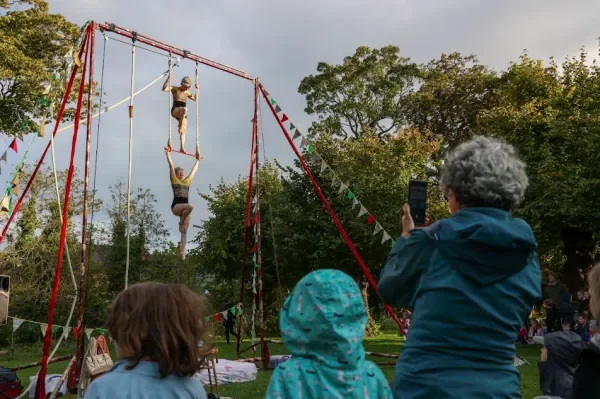Two Aerial Artists perform on red rig in front of a backdrop of green trees while audience look on 