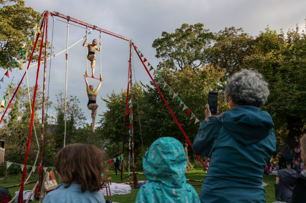 Two Aerial Artists perform on red rig in front of a backdrop of green trees while audience look on 