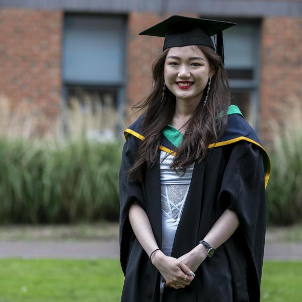 An Asian woman in a graduation cap and gown stands smiling in front of the camera.