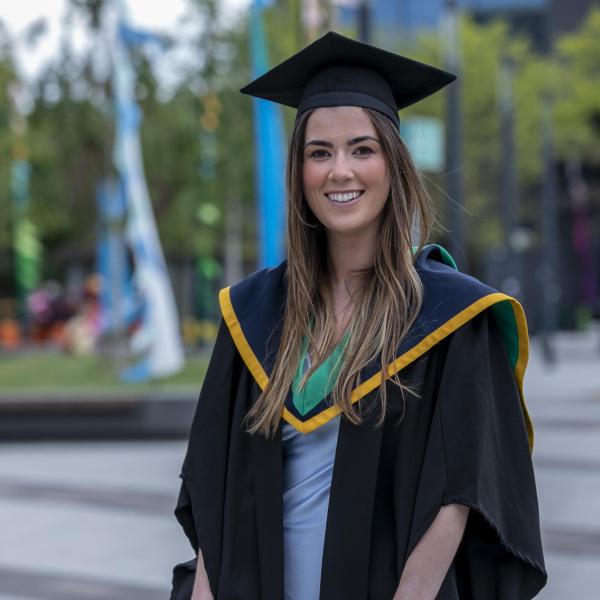 A woman in a graduation cap and gown stands smiling in front of the camera.