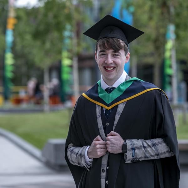 A man in a graduation cap and gown stands smiling widely in front of the camera.