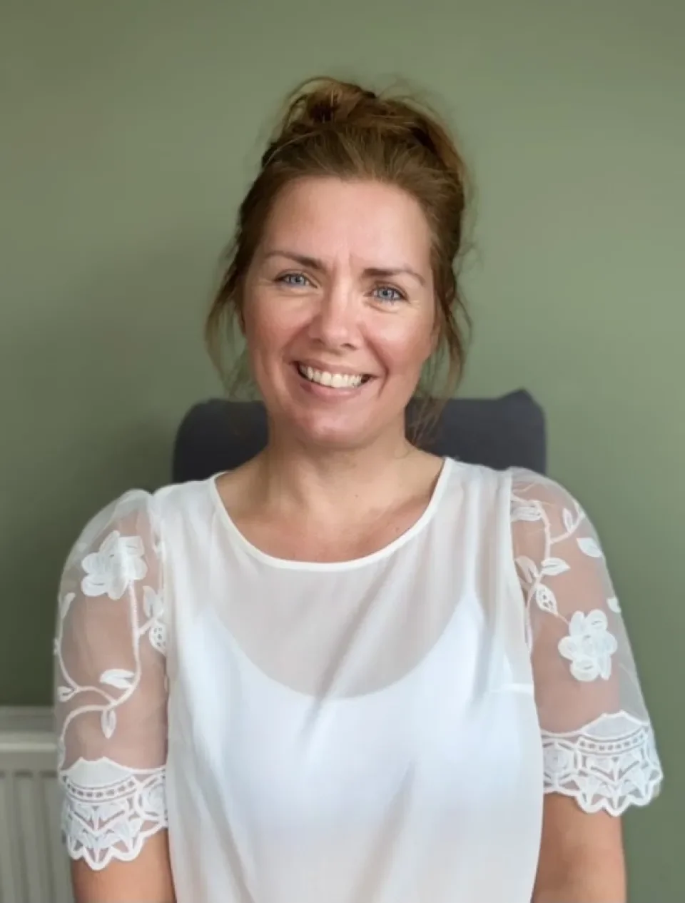 A white woman with brown hair sitting down wearing a white top and smiling at the camera..