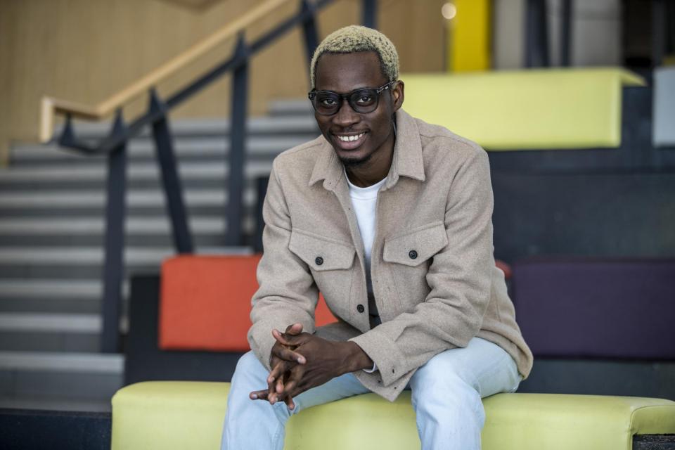 A black man with blonde hair, wearing a beige jacket and light blue jeans sits on a cushioned seat in the U, smiling to the camera.
