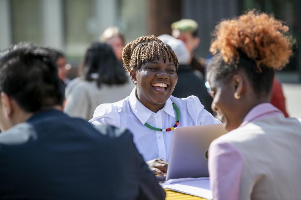 A photo of a black woman sitting in a group outside in the sun. She is wearing a white shirt and has her laptop open in front of her. She is laughing and smiling widely.
