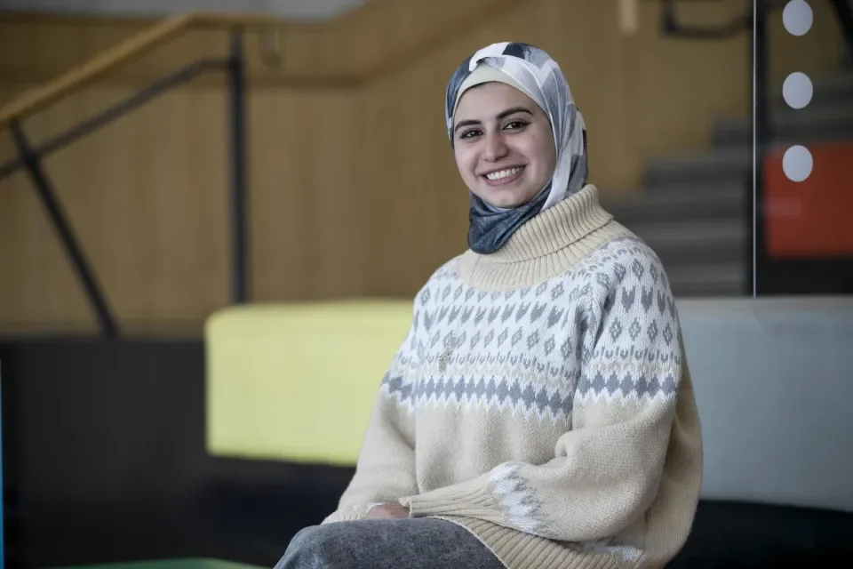 A woman in a headscarf wearing light coloured clothing sits on the coloured steps in the U, smiling to the camera.
