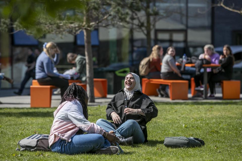 A group of international students sitting outside in the sun on the grass outside the U, smiling and talking to each other.
