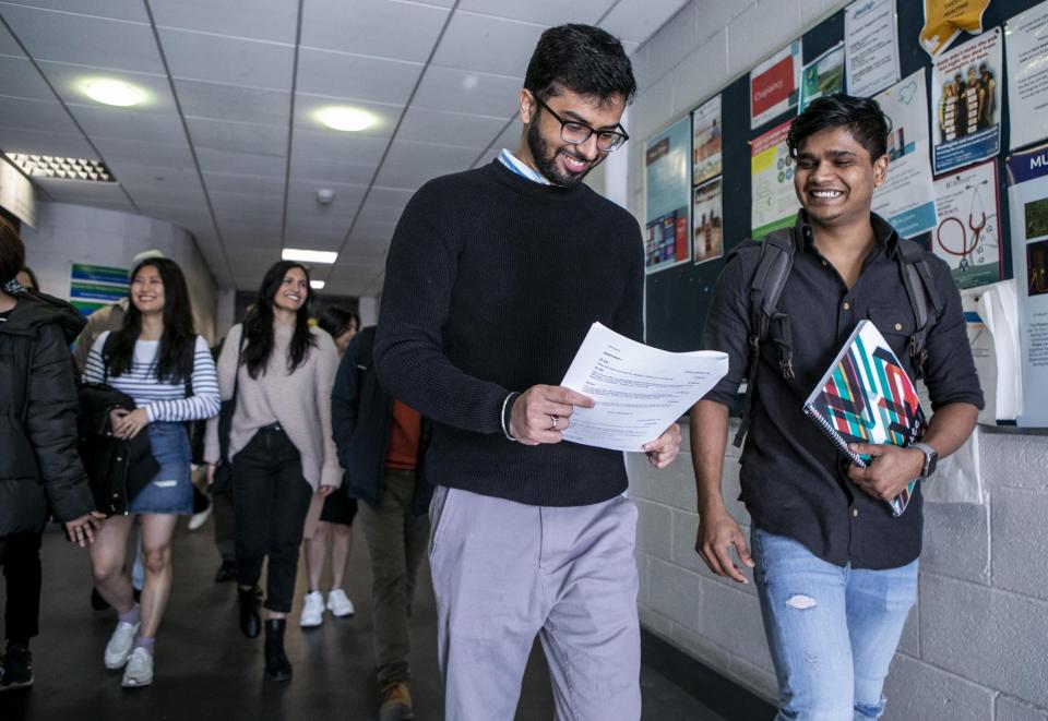 Two Indian men walking ahead of a group in a hallway. Each is holding a piece of paper and one is looking at it, smiling.