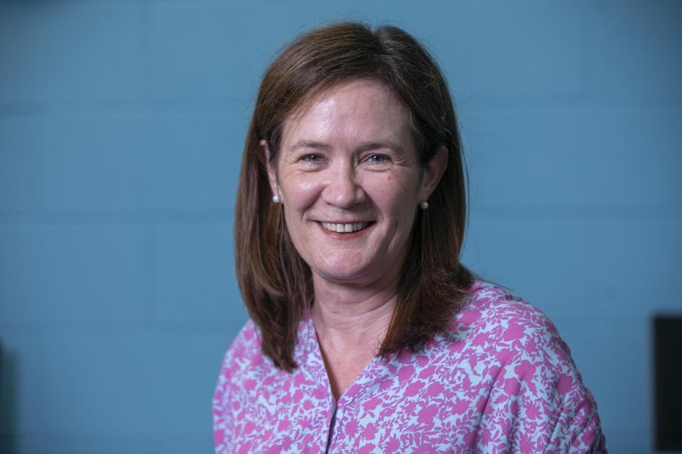 A professional headshot of a woman with brown hair wearing a blue shirt with a purple floral pattern on it. She is smiling into the camera.