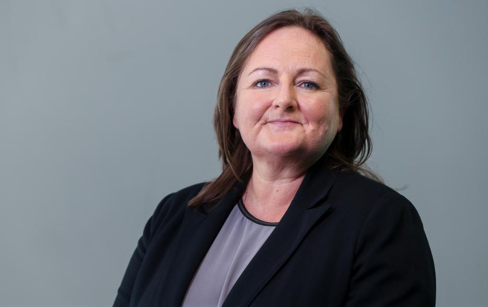 A professional headshot of a middle aged white woman with brown hair wearing a black jacket and a grey shirt.