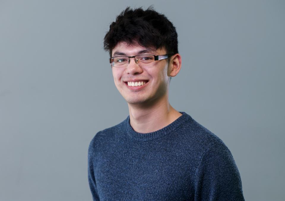 A professional headshot of a man with black hair and brown glasses, wearing a navy jumper, smiling into the camera.