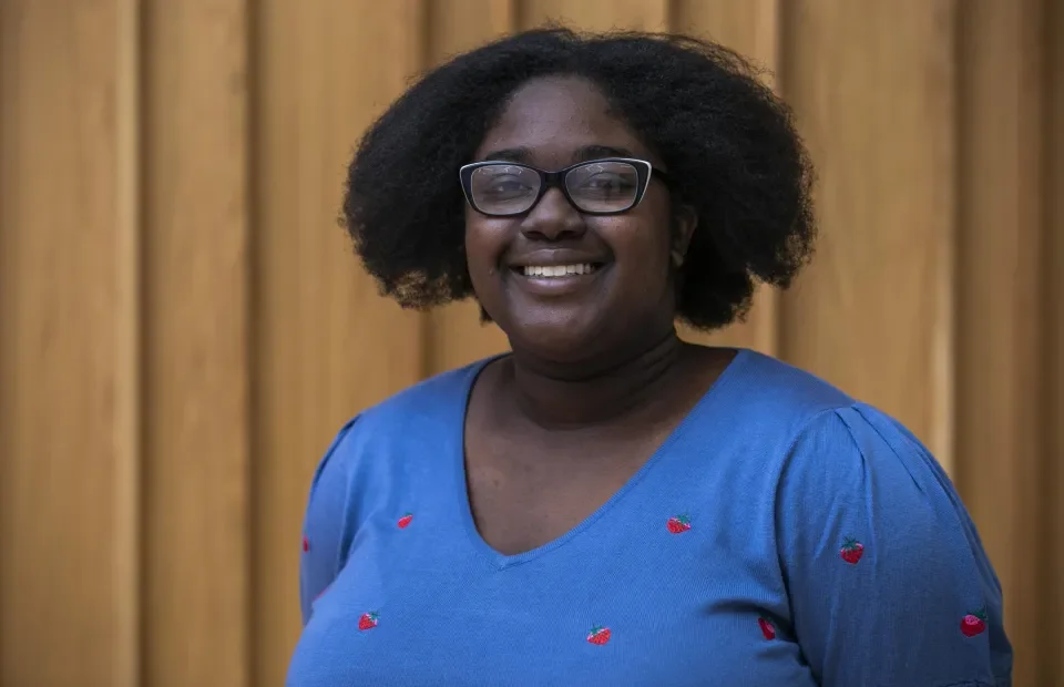 A professional portrait of a black woman with glasses and black hair. She is wearing a blue top with strawberries on it and smiling to the camera.