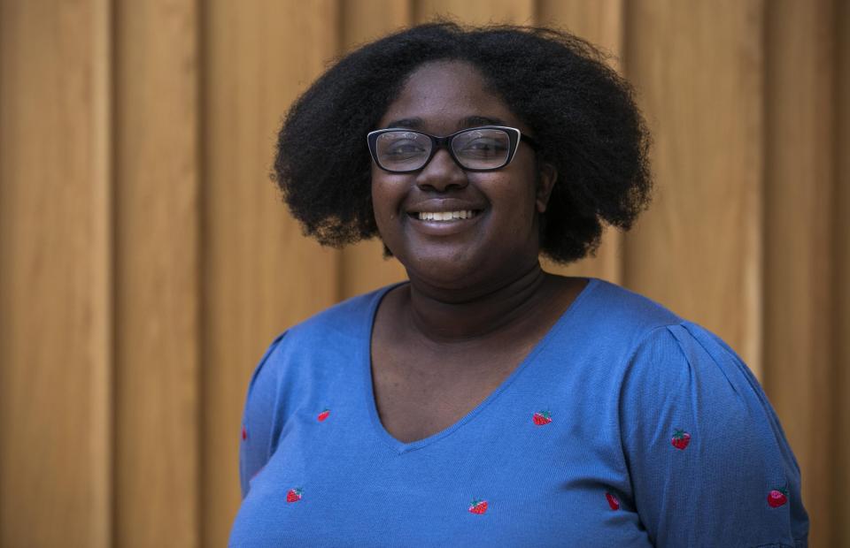 A professional portrait of a black woman with glasses and black hair. She is wearing a blue top with strawberries on it and smiling to the camera. 