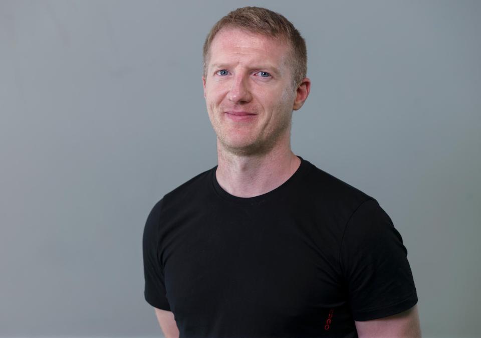 A professional headshot of a white man in a black t-shirt.