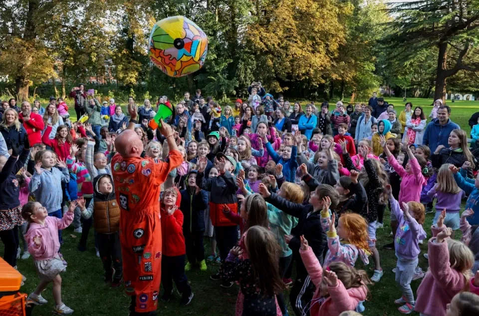 Performer wearing orange jumpsuit throwing large blow up balloon in the air in front of large group or adults and children