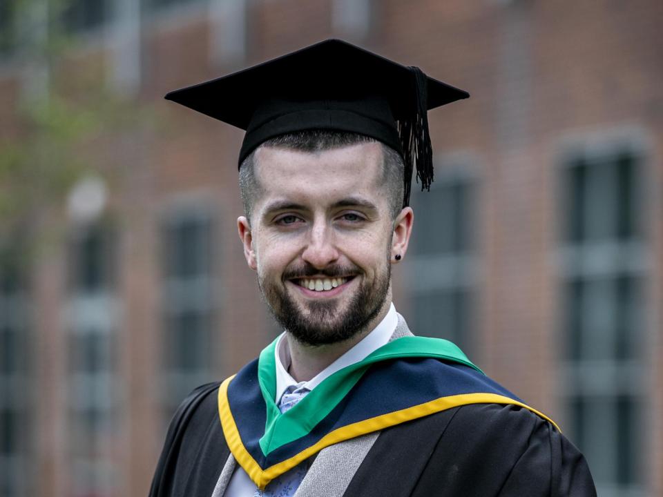 A man poses for the camera, smiling widely, in a graduation cap and gown.