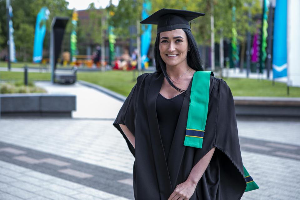 A woman in a graduation cap and gown stands smiling in the middle of the DCU Glasnevin campus.