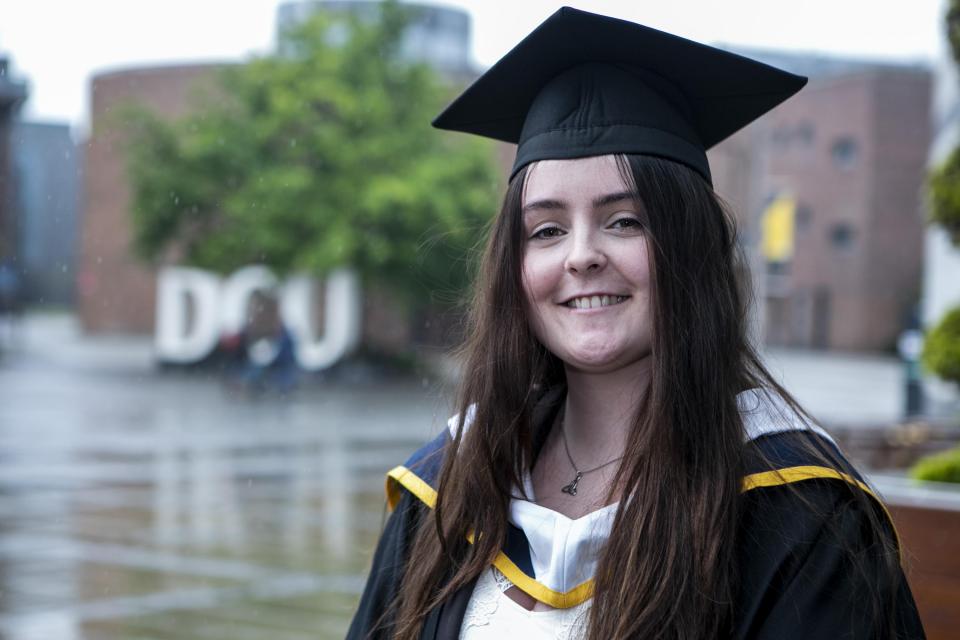 A woman in a graduation cap and gown stands smiling in front of the DCU sign.