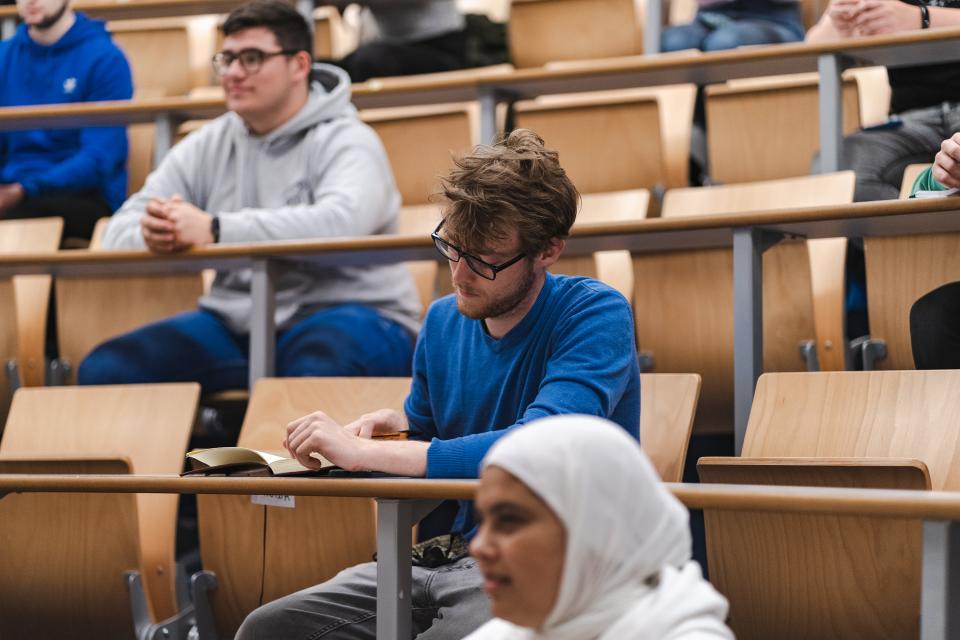 A white man sitting in a lecture hall with other students, taking notes.