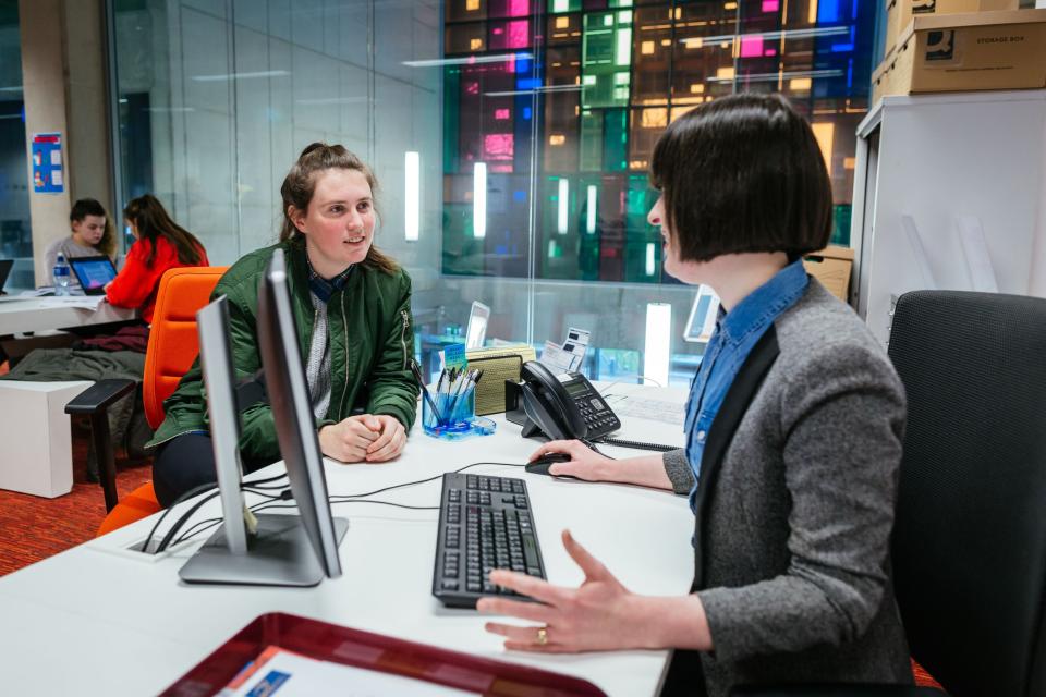 A students sits at a table with a member of library staff, who is giving her information