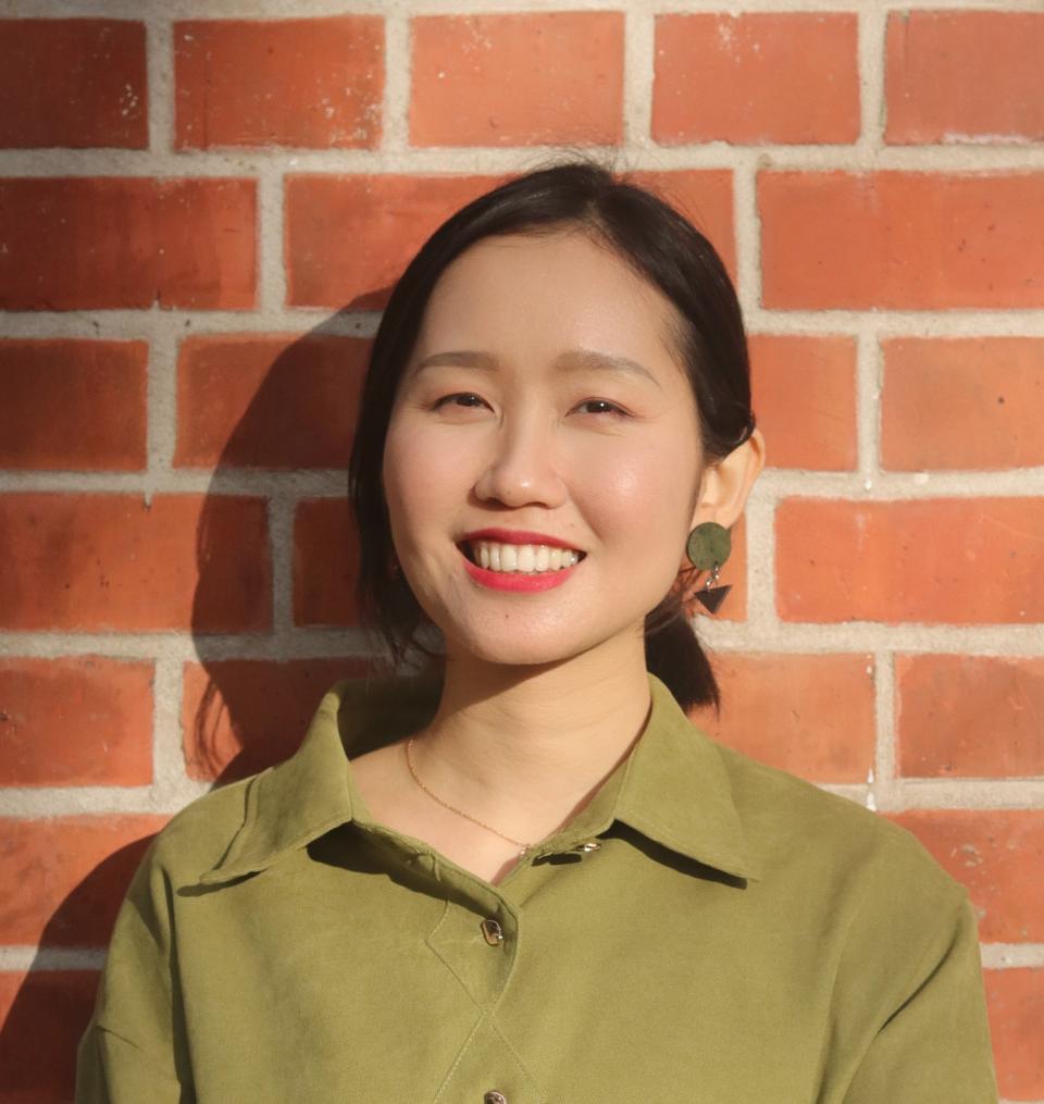 woman with black hair, smiling, in a green shirt against a brick wall