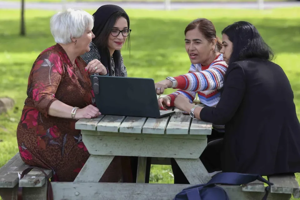 four people sitting down outside chatting while pointing to a laptop