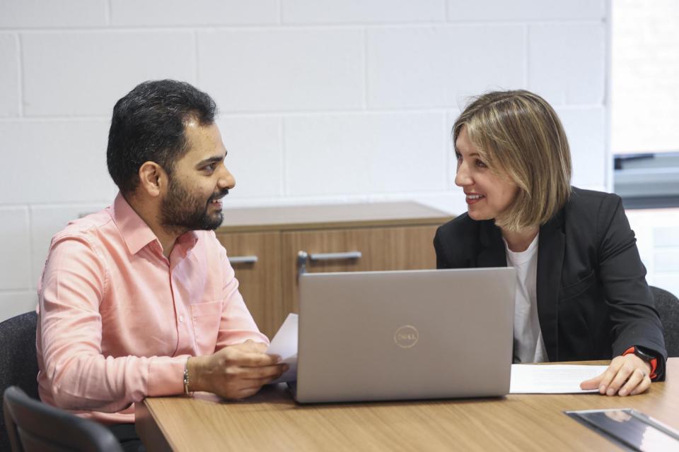 Two people having a meeting in front of a silver laptop. They are smiling and holding sheets of paper.