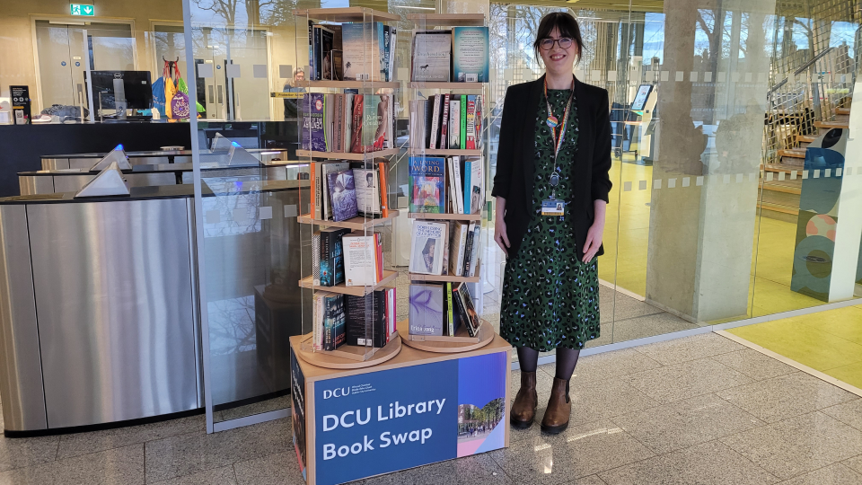 Shows staff member standing beside a carousel filled with books with DCU Library Book Swap written on the stand