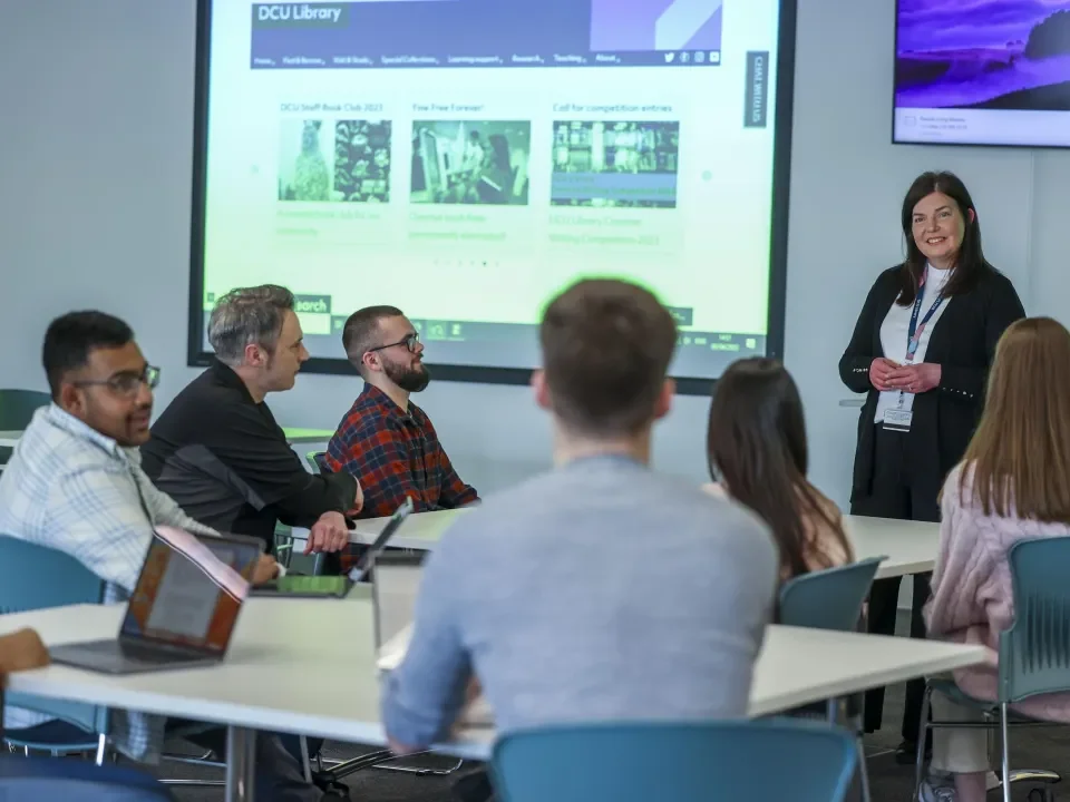 6 students sitting in rows of 3 in a training room, looking at a powerpoint presentation. A librarian stands by the projector, giving a presentation