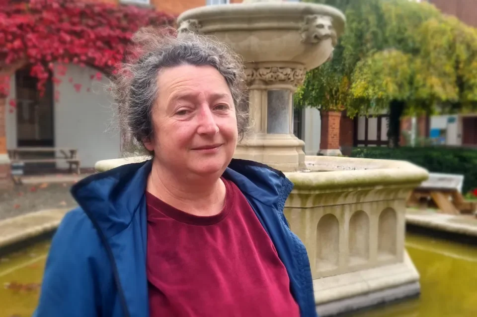 Woman with blue jacket and red jumper standing in front of water fountain 