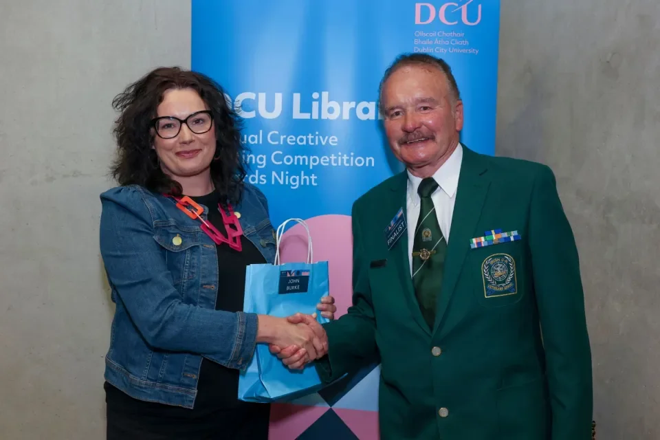 A woman shaking hands with a man, handing him a blue bag. The banner in the background reads DCU Library Annual Creative Writing Competition Awards Night