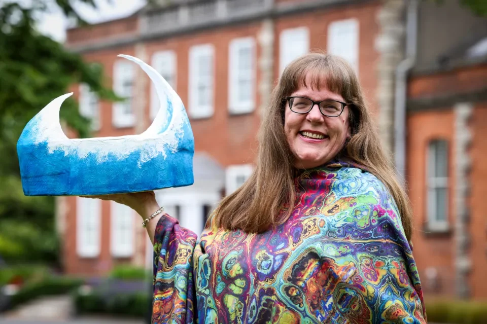 Woman with long hair wearing a colorful tie-dye jacket and holding a handmade papier mache wave