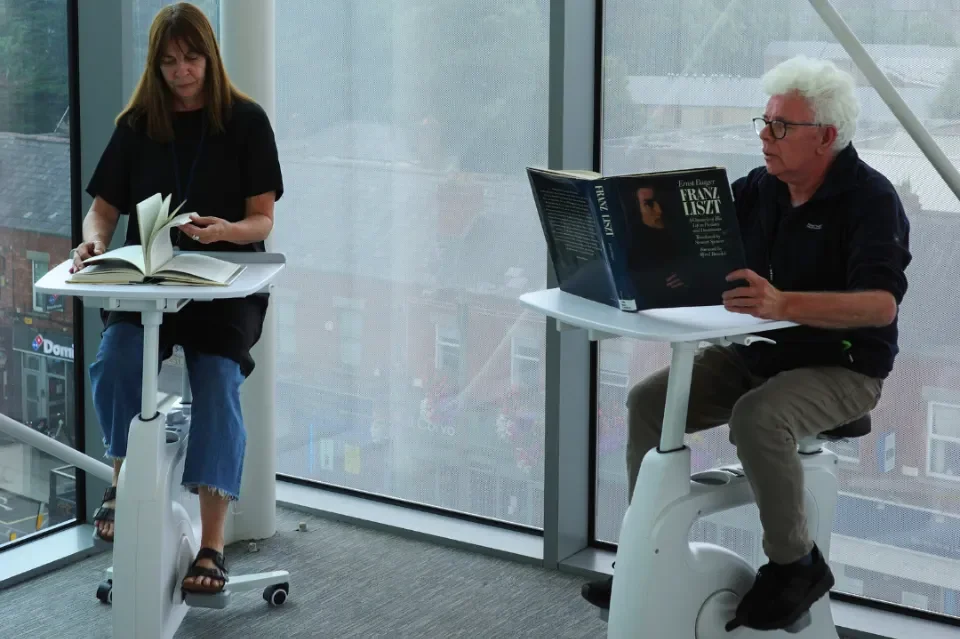 Two people sit on stationary bikes reading books