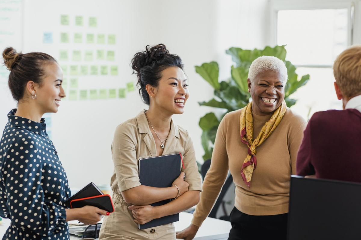 Three women in an office smiling and talking to a male colleague.