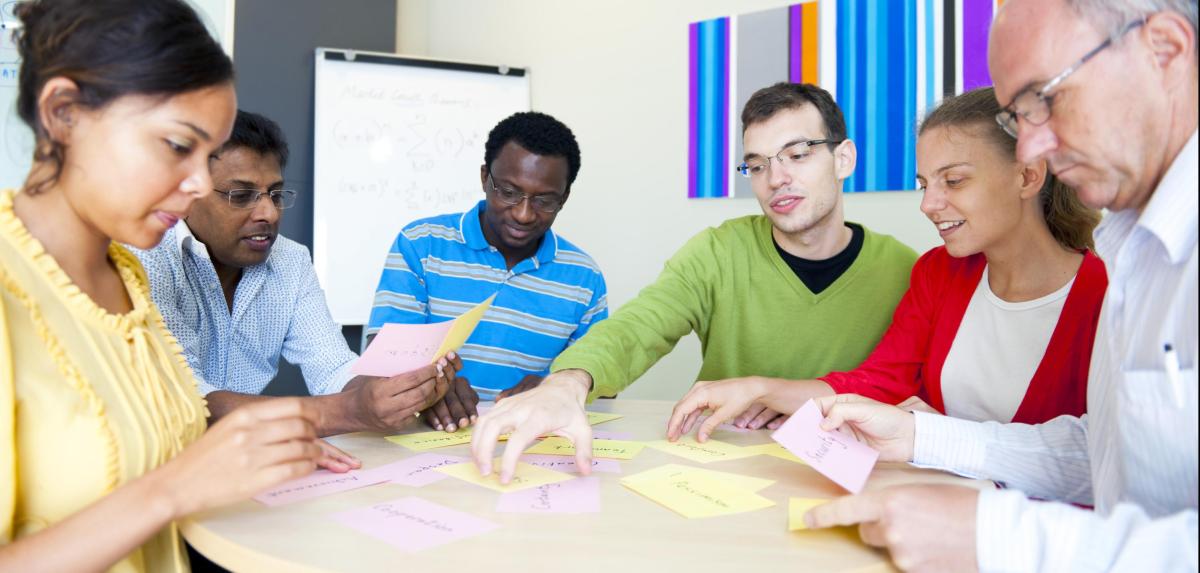 A group of people sitting around a table with colorful paper cards, a whiteboard, and a wall with colorful stripes. Faces are blurred for privacy