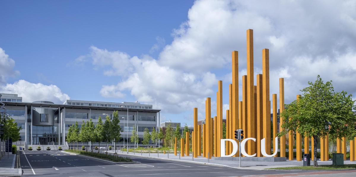 A sunny day in with a modern building on the left and the Orange DCU sticks on the right. the letter DCU have been sculpted in concrete.