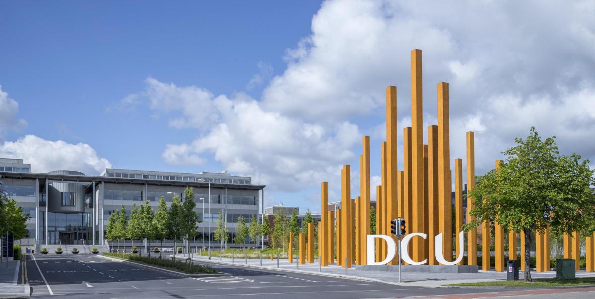 A view of DCU and it's surrounding modern buildings on a sunny day, featuring the DCU sign and the wooden sculpture next to it.